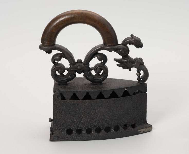 żelazko na węgiel - Ujęcie z boku; Żelazko na węgiel wykonane z żeliwa (odlew). Stopa o kształcie przypominającym trójkąt równoramienny z lekko zaokrąglonymi dłuższymi bokami. Korpus – pojemnik na węgiel – wysoki o prostych ściankach. Wzdłuż jego dolnych, dłuższych krawędzi niewielkie, okrągłe otwory. Klapka przykręcona do korpusu za pomocą śruby. Pokrywa uchylna, mocowana na znajdującym się z tyłu korpusu metalowym pręcie. Do zewnętrznej strony mocowany za pomocą dwóch śrub ozdobny uchwyt. Przednia część uchwytu zdobiona głową smoka jest ruchoma, z rozwidlonym końcem, zaczepianym o mocowany w korpusie pręcik zakończony gałką.