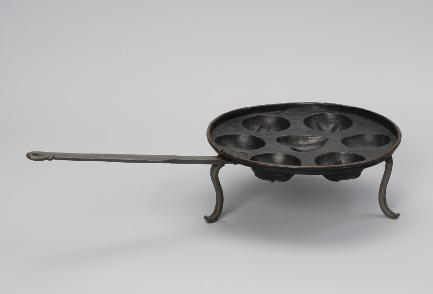 patelnia dołkownica - Ujęcie z boku; Patelnia z miedzianym naczyniem oraz kutymi z żelaza: rączką i trzema nóżkami. Talerz patelni okrągły, miedziany o niskim kołnierzu i z siedmioma wgłębieniami na ciasto. Środkowe wgłębienie w kształcie serca, pozostałe, okalające je – okrągłe. Nóżki dołączone od spodu wykonane z płaskowników, u dołu esowato wygięte na zewnątrz. Przymocowane są do spodniej części dna za pomocą nitów – trzech w każdej nóżce. Do jednej z nóżek mocowana długa, prosta rączka. Na około 2/3 długości spłaszczona. Koniec rączki wygięty esowato i dociśnięty do uchwytu, tworząc otwór w kształcie łezki. Patelnia nosi ślady użytkowania.