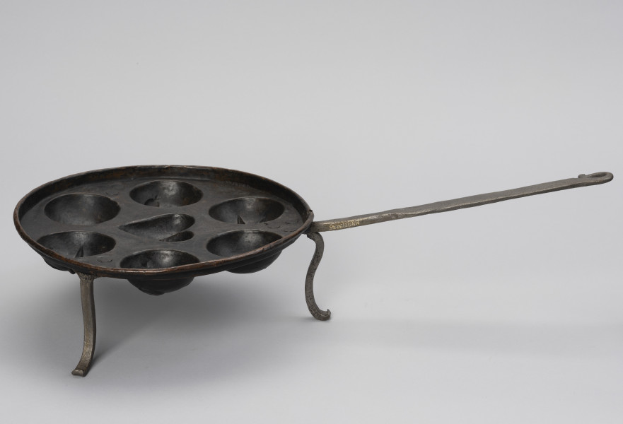 patelnia dołkownica - Ujęcie z boku; Patelnia z miedzianym naczyniem oraz kutymi z żelaza: rączką i nóżkami. Talerz patelni okrągły, miedziany o niskim kołnierzu i z siedmioma wgłębieniami na ciasto. Środkowe wgłębienie w kształcie serca, pozostałe, okalające je – okrągłe. Nóżki dołączone od spodu wykonane z płaskowników, u dołu esowato wygięte na zewnątrz. Przymocowane są do spodniej części dna za pomocą nitów – trzech w każdej nóżce. Do jednej z nóżek mocowana długa, prosta rączka. Na około 2/3 długości spłaszczona. Koniec rączki wygięty esowato i dociśnięty do uchwytu, tworząc otwór w kształcie łezki. Patelnia nosi ślady użytkowania.