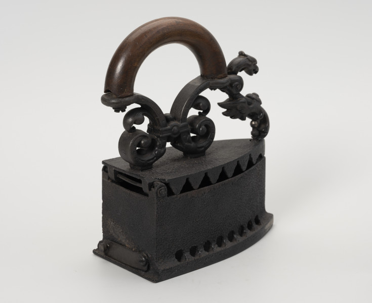 żelazko na węgiel - Ujęcie z tyłu ze skosu z prawej; Żelazko na węgiel wykonane z żeliwa (odlew). Stopa o kształcie przypominającym trójkąt równoramienny z lekko zaokrąglonymi dłuższymi bokami. Korpus – pojemnik na węgiel – wysoki o prostych ściankach. Wzdłuż jego dolnych, dłuższych krawędzi niewielkie, okrągłe otwory. W ściance tylnej prostokątny, wąski otwór zamykany na ruchomą klapkę z niewielką gałką. Klapka przykręcona do korpusu za pomocą śruby. Pokrywa uchylna, mocowana na znajdującym się z tyłu korpusu metalowym pręcie. Do zewnętrznej strony mocowany za pomocą dwóch śrub ozdobny uchwyt. Na jego górną, łukowato wygiętą część nałożona drewniana osłona chroniąca dłoń przed gorącą temperaturą. Przednia część uchwytu zdobiona głową smoka jest ruchoma, z rozwidlonym końcem, zaczepianym o mocowany w korpusie pręcik zakończony gałką.