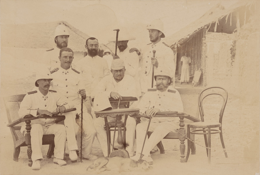 fotografia z albumu Aleksandra Dandelskiego; Aleksander Dandelski w towarzystwie członków ekspedycji wojskowej do Niemieckiej Afryki Wschodniej - ujęcie z przodu; Fotografia w układzie poziomym przedstawia ośmiu mężczyzn ustawionych w dwóch rzędach. W pierwszym czterech z nich siedzi na wyplatanych w stylu kolonialnym fotelach oraz krześle (pierwszy po prawej to Aleksander Dandelski), w drugim kolejnych czterech stoi. Wszyscy ubrani są na biało. Większość ma na sobie mundur, prawdopodobnie oficerski lub elegancki strój. Część z nich ma na głowach białe kolonialne hełmy, a w dłoniach trzyma laski. Przed mężczyznami stoi kawiarniany stolik, na którym postawiono hełm. Po prawej stronie pozostawiono puste krzesło do którego przypięto łańcuchem jeden z foteli. U stóp siedzących mężczyzn leży śpiący pies. W tle widać miejscową kobietę i tubylczą zabudowę.