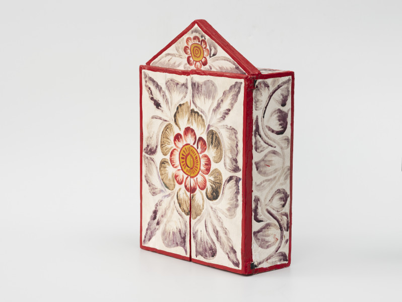 retablo - Ujęcie przodu ze skosu z prawej; Jasna zamknięta, bogato dekorowana kolorowymi wzorami florystycznymi, prostopadłościenna, skrzynka (kapliczka) z dwuskrzydłowymi drzwiczkami, zwieńczona trójkątną koroną.