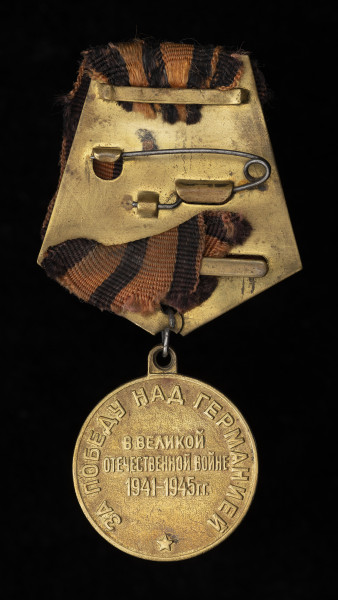 medal Za zwycięstwo nad Niemcami w Wielkiej Wojnie Narodowej 1941-1945 - Ujęcie z tyłu; Medal jest okrągły, z czarno-pomarańczową wstążką na stelażu, z baretką wykonaną z tego samego materiału. Na rewersie widnieją napisy w języku rosyjskim – w otoku: ЗА ПОБЕДУ НАД ГЕРМАНИЕЙ oraz centralnie: В ВЕЛИКОЙ ОТЕЧЕСТВЕННОЙ ВОЙНЕ 1941—1945 ГГ. Poniżej napisu znajduje się pięcioramienna gwiazda.