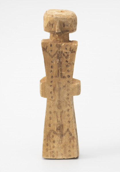figurka magiczna - Ujęcie z przodu; Stojąca figurka postaci ludzkiej o zredukowanej formie wykonana z jasnego drewna. Głowę nieznacznie oddzielono od tułowia wcięciem imitującym szyję. Oczy, usta, nos wyróżniono płytkimi nacięciami oraz czarnym barwnikiem. Na długim, wykonanym schematycznie korpusie w kształcie klepsydry zaznaczono jedynie ręce lub dłonie. Całość pokryto geometrycznymi (linie, łuki, kropki) i antropomorficznymi malunkami wykonanymi wyblakłym już czarnym barwnikiem. Rysunki wykonane na tułowiu ułożono na osi pionowej. Na jasnej powierzchni drewna widoczne są większe i mniejsze spękania, ślady ciosania, malutkie otworki.