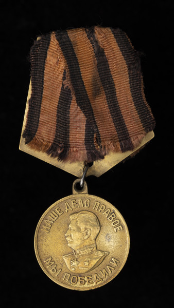 medal Za zwycięstwo nad Niemcami w Wielkiej Wojnie Narodowej 1941-1945 - Ujęcie z przodu; Medal jest okrągły, z czarno-pomarańczową wstążką na stelażu, z baretką wykonaną z tego samego materiału. Na awersie znajduje się wizerunek Józefa Stalina w lewym profilu, otoczony napisem w języku rosyjskim: НАШЕ ДЕЛО ПРАВОЕ | МЫ ПОБЕДИЛИ.