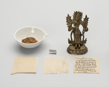 MNS/EP/1077/1-6 figurka Bodhisattwa Awalokiteśwara Padmapani wraz z wypełnieniem - ujęcie z przodu; Z prawej strony mała, wykonana z brązu figurka przedstawiająca Bodhisattwę Awalokiteśwarę Padmapani wraz z wypełnieniem w postaci trzech zwitek papieru, rozłożone na płasko z przodu (dwie zapisane, jedna czysta), z lewej strony figury trzy patyczki (patyczek w trzech częściach? - najprawdopodobniej był to jeden patyczek na który zawinięto arkusze papieru), obok w miseczce materiał sypki na który składa się: zawartość roślinna (ryż, nasiona) oraz glina - przechowywane w osobnej kopercie, kartoniku i woreczku strunowym. Postać ludzka o skupionej, lekko uśmiechniętej twarzy, na głowie diadem. Stoi na lotosowym postumencie o 11 parach odwróconych płatków, którego górną część ozdobiono paciorkami. Ubrana jest w szaty królewskie: przewiązaną w talii tkaninę z paskiem od którego odchodzą łańcuszki z paciorków oraz biżuterię książęcą: trójramienny diadem, parę, dużych, okrągłych kolczyków, podwójny naszyjnik z paciorków z wisiorkami, bransolety na nadgarstkach i przedramionach. Do prawego ramienia przylega kwiat lotosu (typowy atrybut), natomiast po lewej stronie, symetrycznie, na tej samej wysokości, przylega przypominający kwiat lotosu niezidentyfikowany atrybut Awalokiteśwary. Obie dłonie ułożone są w geście argumentacji: kciuk i palec wskazujący złączone, tworzą krąg. Prawa uniesiona jest na wysokości klatki piersiowej, lewa opada wzdłuż ciała, zatrzymując się na wysokości biodra. Z ramion figurki zwisa przewieszona skóra antylopy.