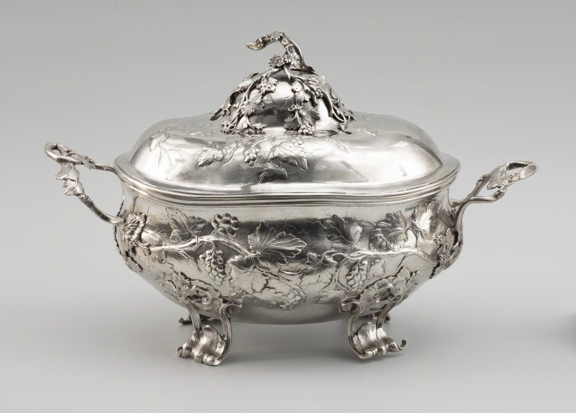 waza - Ujęcie z przodu; Owalna teryna, wsparta na czterech nóżkach. Korpus o płynnym, organicznym kształcie i falistej linii brzegu, w dolnej części wybrzuszony. Pokrywa wysoka, dwustopniowo wybrzuszona. Zarówno korpus, jak i pokrywa zdobione repusowanymi w płytkim reliefie naturalistycznymi gałązkami winorośli z gronami owoców. Reliefowy ornament miękko przechodzi w wykonane techniką odlewu, plastyczne gałązki winnej latorośli, tworzące dwa uchwyty przy korpusie oraz ozdobne zwieńczenie pokrywy. Nóżki wolutowo wygięte, ażurowe, zdobione rocaillami i gałązkami winorośli.