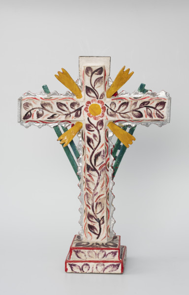 krzyż pasyjny - Ujęcie z tyłu; Drewniany, czerwony krzyż pasyjny, usytuowany na białej, dwustopniowej, kwadratowej podstawie ozdobionej ciemnym malowanym wzorem półkolistych form i liści, której krawędzie pokryto czerwoną farbą. Bogato dekorowany białymi i fioletowymi wzorami kwiatowymi i obłymi liniami, dodatkowo boki ramion krzyża zostały ozdobione plastycznym falistym motywem w kolorze srebrnym, którego krawędzie podkreślono czerwoną farbą. Narożniki między skrzyżowanymi ramionami krucyfiksu udekorowano czterema potrójnymi promieniami w kolorze pomarańczowym. Całość pokryto białą masą i malowanym wzorem florystycznym (pojedyncza, ciemna, liściasta wić pokrywająca ramiona krzyża oraz żółto-czerwony kwiat w miejscu przecięcia ramion – stosowanym również w skrzyneczkach retablos).