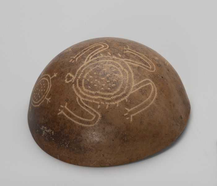 Naczynie do serwowania jedzenia, napojów i guarapo - Ujęcie z przodu, spód misy; Miska z tykwy o naturalnym jasnobrązowym, orzechowym kolorze z zewnętrzną powierzchnią bogato zdobioną rytymi ornamentami zoomorficznymi i jednym antropomorficznym. Ornamentacja wykonywana za pomocą metalowego narzędzia, jego ślady w postaci płytkich i drobnych nacięć widoczne są na wyrytych rysunkach przedstawiających trudne do zidentyfikowania zwierzęta. Największy z nich to najprawdopodobniej żółw lądowy w towarzystwie mniejszego żółwia. Na zewnętrznej powierzchni tykwy widoczne są mniejsze nacięcia i wgłębienia, przetarcia oraz ślady użytkowania.