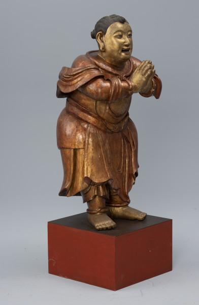 pielgrzym Shancai Tongzi (?) wraz z wypełnieniem - ujęcie z przodu z lewej; Monumentalna, drewniana, złocona rzeźba postaci męskiej w pozie adoracji przedstawia prawdopodobnie młodego pielgrzyma. Postać stoi na postumencie z rękami wyciągniętymi przed siebie i dłońmi złożonymi w pełnym szacunku geście. Ma pogodny wyraz twarzy, lekko uniesione brwi, duże, brązowe oczy, podłużne uszy i otwarte, lekko uśmiechnięte usta z widocznym uzębieniem. W jego policzkach zaznaczono dołki, natomiast w płatkach usznych wydrążono niewielkie otwory. Gładko zaczesane czarne włosy zwijają się w niewielkie koczki: z przodu jeden i z tyłu dwa. Pielgrzym ubrany jest w długie, kilkowarstwowe, sięgające za kolana szaty, natomiast z jego ramion i rąk opada tkanina w formie szala. Na jego obu nadgarstkach i kostkach widoczne są bransolety. Stopy ma nagie.