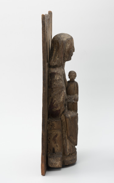 Tronująca Madonna z Dzieciątkiem - Ujęcie z lewej; Niewielka figura tronującej Marii z dzieciątkiem o zwartej bryle, przytwierdzona do wysokiego zaplecka o geometrycznym zwieńczeniu, polichromowana. Rzeźba opracowana jako przyścienna. Brak prawej dłoni Marii. Powierzchniowe ubytki drewna i otwory po drewnojadach. Polichromia zachowana szczątkowo.