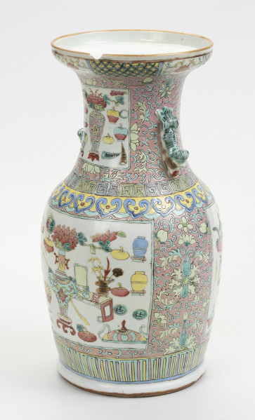 wazon typu famille rose - ujęcie z przodu z prawej; Porcelanowy wazon zdobiony wielobarwną dekoracją naszkliwną w pastelowych odcieniach z przewagą różu w typie famille rose. Kształt gruszkowaty, szyja szeroka zakończona rozszerzającym się wylewem, w którym widoczny jest z przodu półokrągły ubytek. Wazon bogato zdobiony licznymi motywami dekoracyjnymi. W jego górnej części z prawej znajduje się uchwyt w formie stylizowanego lwa chińskiego w kolorze pastelowej zieleni. Brzeg wylewu zdobiony dookolnie i naprzemiennie motywami stylizowanego lotosu o czerwonym kwiecie, żółtych i zielonych liściach na białym tle oraz zgeometryzowanym wzorem roślinnym w kolorach zielonym i różowym na żółtym tle. Szyję oddzielono od brzuśca biegnącymi dookolnie wzorami: ornamentem meandrycznym o różowej, żółtej i seledynowej barwie oraz znajdującym się pod nim motywem grzyba nieśmiertelności w niebieskim, żółtym, czerwonym i seledynowym kolorze. Brzusiec oraz szyję zdobią czworoboczne pola (rezerwy) o seledynowych bordiurach wypełnione tzw. motywem Stu Antyków (Hundred Antiques/Hundred Treasures) na białym tle. Pola wypełniają przedmioty i rośliny w dość nieformalnym układzie. Na szyi wyodrębnić można: rogi nosorożca, śliwki, wazon z peoniami na stojaku, różnorodne wazy (z przykrywkami oraz inne niezidentyfikowane przedmioty. Na brzuścu pokazano: śliwki, wazę z kwiatami na stojaku,  toaletkę z lustrem, różnorodne wazy, oraz inne niezidentyfikowane przedmioty i rośliny. Tło stanowi przedstawiony na różowym tle motyw stylizowanego lotosu z wijącymi się liśćmi. Dół wazonu ozdobiony dookolnie geometrycznym motywem w postaci pionowych żółtych i różowych pasków na seledynowym tle.