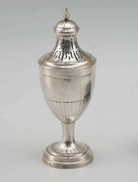 cukiernica do cukru pudru - Ujęcie z przodu; Puszka z ażurową pokrywą, kształtem nawiązująca do antycznych urn. Wsparta na okrągłej stopie z trzema uskokami, przechodzącej w niewysoki, drobno kanelowany trzon. Korpus smukły, o kielichowej formie, w dolnej części również kanelowany. Pokrywa wysoka, o wklęsłych ściankach zakończona półkolistą kopułką, ze zwieńczeniem w formie maleńkiej wazy. Wycięcia na pokrywie układają się we wzór stylizowanych kampanul.