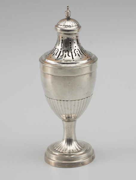 cukiernica do cukru pudru - Ujęcie z przodu; Puszka z ażurową pokrywą, kształtem nawiązująca do antycznych urn. Wsparta na okrągłej stopie z trzema uskokami, przechodzącej w niewysoki, drobno kanelowany trzon. Korpus smukły, o kielichowej formie, w dolnej części również kanelowany. Pokrywa wysoka, o wklęsłych ściankach zakończona półkolistą kopułką, ze zwieńczeniem w formie maleńkiej wazy. Wycięcia na pokrywie układają się we wzór stylizowanych kampanul.