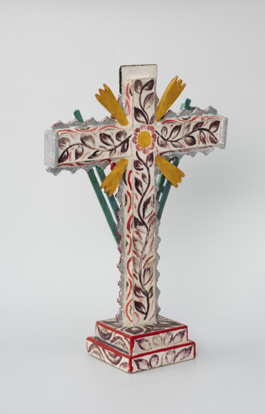 krzyż pasyjny - Ujęcie z tyłu z lewej; Drewniany, czerwony krzyż pasyjny, usytuowany na białej, dwustopniowej, kwadratowej podstawie ozdobionej ciemnym malowanym wzorem półkolistych form i liści, której krawędzie pokryto czerwoną farbą. Bogato dekorowany białymi i fioletowymi wzorami kwiatowymi i obłymi liniami, dodatkowo boki ramion krzyża zostały ozdobione plastycznym falistym motywem w kolorze srebrnym, którego krawędzie podkreślono czerwoną farbą. Narożniki między skrzyżowanymi ramionami krucyfiksu udekorowano czterema potrójnymi promieniami w kolorze pomarańczowym. Całość pokryto białą masą i malowanym wzorem florystycznym (pojedyncza, ciemna, liściasta wić pokrywająca ramiona krzyża oraz żółto-czerwony kwiat w miejscu przecięcia ramion – stosowanym również w skrzyneczkach retablos).