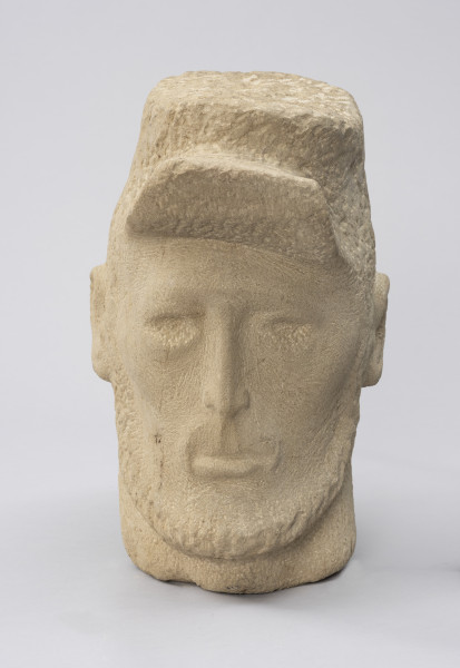 rzeźba; głowa; Komunard - ujęcie z przodu; Głowa męska w czapce od uniformu Gwardii Narodowej z uniesionym daszkiem. Pociągła twarz o delikatnie zaznaczonych rysach uformowana przez minimalistyczne opracowanie jasnego, kamiennego surowca. Jedno oko przymrużone, nos długi i szczupły, wokół ust o grubej dolnej wardze delikatny wąsik, zarost zaznaczony też wzdłuż żuchwy, szyja szeroka. Pozostawione na powierzchni czapki i brody ślady pracy grubym dłutem dają wrażenie miękkości. Zmarszczki ogorzałej skóry mężczyzny uzyskano dzięki pracy cieńszymi dłutami.