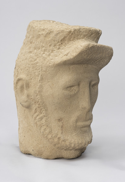 rzeźba; głowa; Komunard - ujęcie ze skosu z lewej; Głowa męska w czapce od uniformu Gwardii Narodowej z uniesionym daszkiem. Pociągła twarz o delikatnie zaznaczonych rysach uformowana przez minimalistyczne opracowanie jasnego, kamiennego surowca. Jedno oko przymrużone, nos długi i szczupły, wokół ust o grubej dolnej wardze delikatny wąsik, zarost zaznaczony też wzdłuż żuchwy, szyja szeroka. Pozostawione na powierzchni czapki i brody ślady pracy grubym dłutem dają wrażenie miękkości. Zmarszczki ogorzałej skóry mężczyzny uzyskano dzięki pracy cieńszymi dłutami.