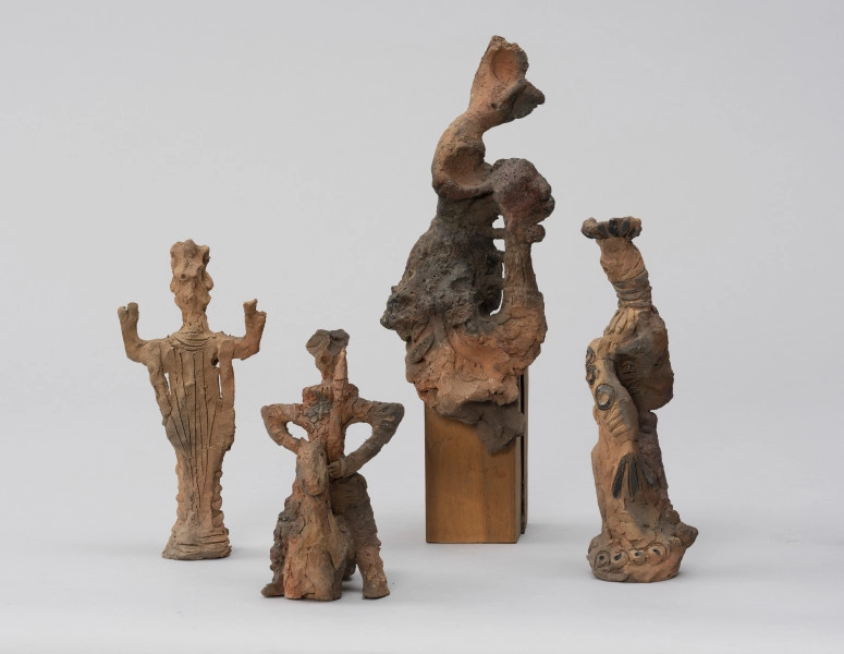 zespół rzeźb; Zmierzch królów - ujęcie z przodu; Zespół surrealistycznych antropomorficznych i zoomorficznych figur o ceglasto-szarej barwie, od lewej:
a) Postać męska o zwartych nogach, uniesionych w górę, zgiętych w łokciach ramionach. Sterczące nakrycie głowy, spodnie bufiaste na biodrach, na nogach poprzecznie nałożone paski gliny. Na torsie i szyi ryty.
b) Jeździec na koniu bez nóg. Szeroko rozstawione ręce, wsparte dłońmi o biodra, w lewej sterczący w górę miecz. Głowa mała na długiej szyi, na szyi kryza. Na torsie rytem zaznaczone łuski pancerza. W stosunku do postaci koń mały, schematycznie potraktowany.
c) Ogólnie potraktowanej stojącej na postumencie postaci kobiecej. Głowa odchylona w prawo, w tył, tors w lewo, fragmentarycznie zaznaczony. Od bioder draperie fałd spódnicy. Po prawej, przy postaci pionowa forma połączona trzema poziomymi trzpieniami z postacią. Na wysokości torsu postaci forma nieco powiększa się i łączy z postacią.
d) Skręcona spiralnie, wysmukła postać kobieca o tektonicznej formie. Twarz pociągła, na głowie duży, płaski beret. Na grubej szyi rytmiczne krążki. Prawe ramię opuszczone ukośnie w dół, w ręce wachlarz. Na ramieniu trzy czarne krążki. Na podstawie 4 