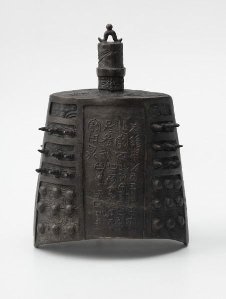 zhong (dzwon chiński) - ujęcie z przodu; Wykonany z brązu dzwon o soczewkowatym przekroju poprzecznym. Na płaskim hełmie znajduje się korona w formie trzpienia, zdobionego rysunkami ptaków, z małym pałąkowatym uszkiem na szczycie zwieńczonym kuleczką i drugim, wygiętym pod kątem prostym, wychodzącym z hełmu i połączonym z trzpieniem za pomocą okrągłej obejmy. Na bogaty ornament reliefowy w postaci siatki wzoru geometrycznego (meander) i zoomorficznego (przedstawienia wężowatych stworzeń, być może smoków) po bokach nałożono kolce mei (w górnej części) oraz guzki (w dolnej części), natomiast w centrum płaszcza wykonano inskrypcję chińskim pismem małopieczęciowym. Dzwon tego typu nie posiada serca, jego środek pozostaje pusty.