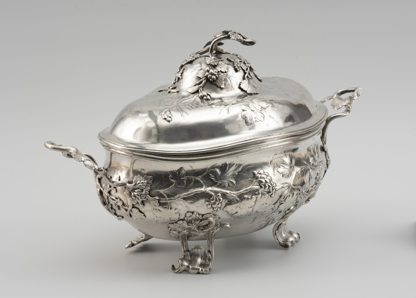 waza - Ujęcie tyłu ze skosu z lewej; Owalna teryna, wsparta na czterech nóżkach. Korpus o płynnym, organicznym kształcie i falistej linii brzegu, w dolnej części wybrzuszony. Pokrywa wysoka, dwustopniowo wybrzuszona. Zarówno korpus, jak i pokrywa zdobione repusowanymi w płytkim reliefie naturalistycznymi gałązkami winorośli z gronami owoców. Reliefowy ornament miękko przechodzi w wykonane techniką odlewu, plastyczne gałązki winnej latorośli, tworzące dwa uchwyty przy korpusie oraz ozdobne zwieńczenie pokrywy. Nóżki wolutowo wygięte, ażurowe, zdobione rocaillami i gałązkami winorośli.