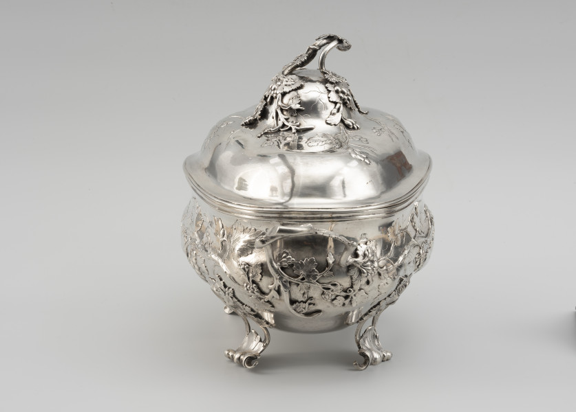 waza - Ujęcie z prawej; Owalna teryna, wsparta na nóżkach. Korpus o płynnym, organicznym kształcie i falistej linii brzegu, w dolnej części wybrzuszony. Pokrywa wysoka, dwustopniowo wybrzuszona. Zarówno korpus, jak i pokrywa zdobione repusowanymi w płytkim reliefie naturalistycznymi gałązkami winorośli z gronami owoców. Reliefowy ornament miękko przechodzi w wykonane techniką odlewu, plastyczne gałązki winnej latorośli, tworzące dwa uchwyty przy korpusie oraz ozdobne zwieńczenie pokrywy. Nóżki wolutowo wygięte, ażurowe, zdobione rocaillami i gałązkami winorośli.