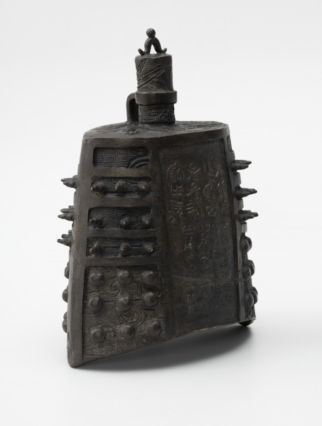 zhong (dzwon chiński) - ujęcie przodu ze skosu z lewej; Wykonany z brązu dzwon o soczewkowatym przekroju poprzecznym. Na płaskim hełmie znajduje się korona w formie trzpienia, zdobionego rysunkami ptaków, z małym pałąkowatym uszkiem na szczycie zwieńczonym kuleczką i drugim, wygiętym pod kątem prostym, wychodzącym z hełmu i połączonym z trzpieniem za pomocą okrągłej obejmy. Na bogaty ornament reliefowy w postaci siatki wzoru geometrycznego (meander) i zoomorficznego (przedstawienia wężowatych stworzeń, być może smoków) po bokach nałożono kolce mei (w górnej części) oraz guzki (w dolnej części), natomiast w centrum płaszcza wykonano inskrypcję chińskim pismem małopieczęciowym.