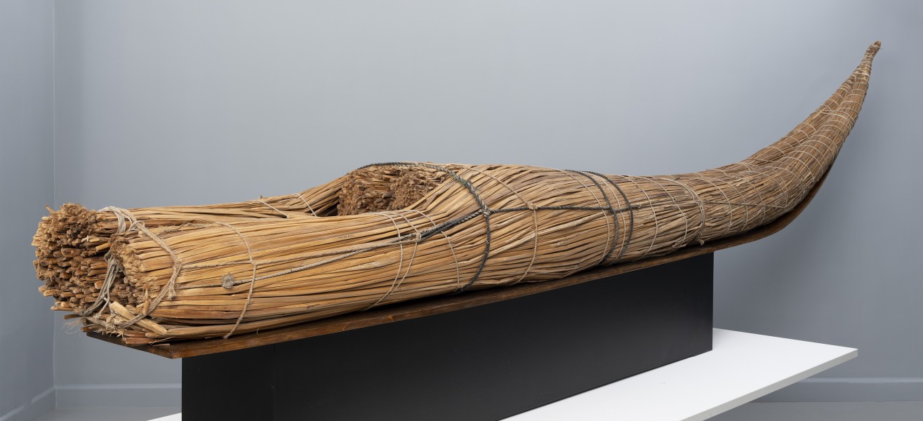 caballito de totora (łódź trzcinowa) - Ujęcie tyłu ze skosu z prawej; Ponad czterometrowa łódź trzcinowa o naturalnym jasnobrązowym kolorze z wysoko zadartym ku górze długim, zwężającym się dziobem oraz grubą, płasko ściętą rufą. Kształtem przypomina gruby cios słonia. Została zbudowana z dwóch wiązek trzciny mocno skrępowanych przez poprzeczne wiązania. W ten sposób nadano łodzi pożądany, żeglowny kształt.