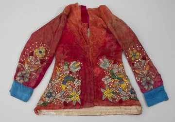 kaftan do tańca negritos - Ujęcie z przodu; Czerwony, lekko wyblakły, welurowy kaftan o trapezowatym kształcie i luźnym kroju, bogato haftowany, ozdobiony kolorowymi cekinami i szklanymi paciorkami. Na rękawach i przedniej części dominują wzory florystyczne, po lewej stronie wyhaftowano kolorowego ptaka przypominającego papugę. Rękawy wykończono niebieską satyną, na welurowej tkaninie zauważalne odbijające się wzory kwiatowe. Na czerwonym welurze miejscami widoczne pozostałości odciskanego wzoru kwiatowego - szczególnie widoczne przy dekolcie. Widoczne naprawy - ślady szycia.