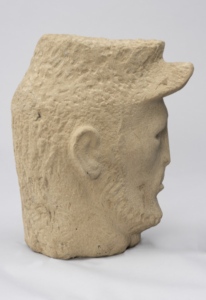 rzeźba; głowa; Komunard - ujęcie z lewej; Głowa męska w czapce od uniformu Gwardii Narodowej z uniesionym daszkiem. Pociągła twarz o delikatnie zaznaczonych rysach uformowana przez minimalistyczne opracowanie jasnego, kamiennego surowca. Nos długi, szyja szeroka. Pozostawione na powierzchni czapki i brody ślady pracy grubym dłutem dają wrażenie miękkości. Zmarszczki ogorzałej skóry mężczyzny uzyskano dzięki pracy cieńszymi dłutami.