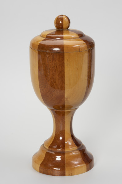 puchar z pokrywką - ujęcie z przodu; Pamiątkowy drewniany puchar ze zdejmowaną pokrywką, zakończoną okrągłym uchwytem. Obrobiony na tokarce z drewnianego klocka klejonego z koncentrycznie ułożonych kawałków drewna w dwóch odcieniach – jasno-żółtawym i ciemno-brązowym – ułożonych naprzemiennie. Pokrywa uformowana na wzór wycinka spłaszczonej, podwójnej bańki z gałką na szczycie. Czara o formie niewysokiego cylindra o zaokrąglonym dnie osadzona jest na kolumnowej, przewężonej nóżce o stopie w formie ściętego stożka.