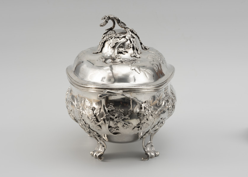 waza - Ujęcie z lewej; Owalna teryna, wsparta na nóżkach. Korpus o płynnym, organicznym kształcie i falistej linii brzegu, w dolnej części wybrzuszony. Pokrywa wysoka, dwustopniowo wybrzuszona. Zarówno korpus, jak i pokrywa zdobione repusowanymi w płytkim reliefie naturalistycznymi gałązkami winorośli z gronami owoców. Reliefowy ornament miękko przechodzi w wykonane techniką odlewu, plastyczne gałązki winnej latorośli, tworzące dwa uchwyty przy korpusie oraz ozdobne zwieńczenie pokrywy. Nóżki wolutowo wygięte, ażurowe, zdobione rocaillami i gałązkami winorośli.