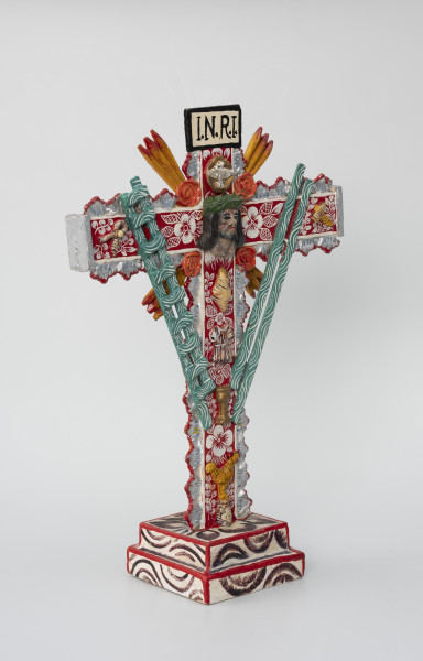 krzyż pasyjny - Ujęcie ze skosu z lewej; Drewniany, czerwony krzyż pasyjny, usytuowany na białej, dwustopniowej, kwadratowej podstawie ozdobionej ciemnym malowanym wzorem półkolistych form i liści, której krawędzie pokryto czerwoną farbą. Bogato dekorowany białymi i fioletowymi wzorami kwiatowymi i obłymi liniami, dodatkowo boki ramion krzyża zostały ozdobione plastycznym falistym motywem w kolorze srebrnym, którego krawędzie podkreślono czerwoną farbą. W jego centralnej części umieszczono wykonaną z formy głowę cierpiącego Chrystusa w koronie cierniowej, nad nią zaś postać gołębia i napisane na prostokątnej plakietce czarnym tuszem litery I.N.R.I. Całość wypełniono rozlicznymi atrybutami i symbolami męki pańskiej, takimi jak: serce, tunika, kielich, kogut, zielona drabina, zielone włócznie (?), szczypce i młotek. Narożniki między skrzyżowanymi ramionami krucyfiksu udekorowano czterema koralowymi różami oraz czterema potrójnymi promieniami w kolorze pomarańczowym.