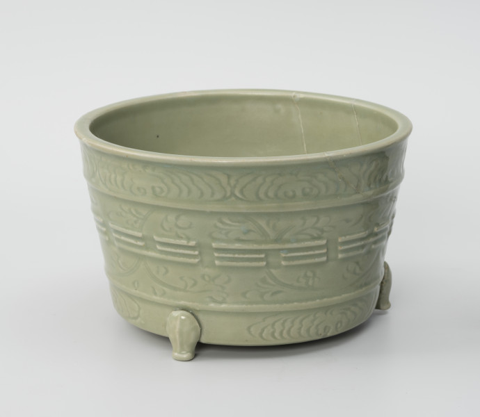 donica ozdobna - Ujęcie z przodu; Kamionkowa donica z trzema dekoracyjnymi nóżkami , widoczne dwie z trzechs, zkliwiona celadonem w jasnozielonym odcieniu. Ma kształt odwróconego, ściętego stożka. Jej nogi stylizowane są na łapy zwierzęce, zostały doklejone do wykonanego na kole garncarskim korpusu. Powierzchnia naczynia została ozdobiona wklęsło-wypukłym ornamentem reliefowym, podzielonym na trzy poziome strefy oddzielone dookolnie biegnącymi listwami. Górny i dolny pas wypełniono motywami roślinnymi – najprawdopodobniej peoniami, środkowa część natomiast składa się ze wzoru roślinnego (peonie) oraz wypukłych linii ułożonych w różnych sekwencjach, przedstawiających osiem podstawowych trygramów. Na ściance z prawej widoczne ślady klejenia.