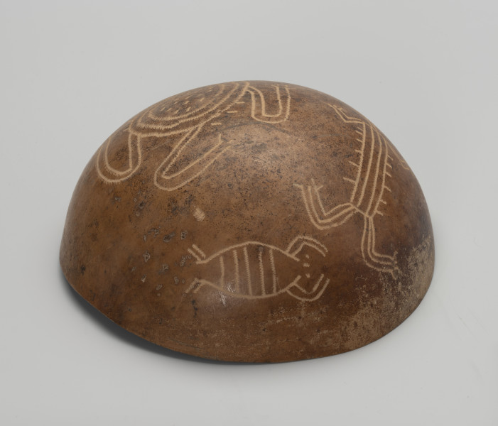 Naczynie do serwowania jedzenia, napojów i guarapo - Ujęcie spodu misy; Miska z tykwy o naturalnym jasnobrązowym, orzechowym kolorze z zewnętrzną powierzchnią bogato zdobioną rytymi ornamentami zoomorficznymi i jednym antropomorficznym. Ornamentacja wykonywana za pomocą metalowego narzędzia, jego ślady w postaci płytkich i drobnych nacięć widoczne są na wyrytych czterech rysunkach przedstawiających postać ludzką oraz trudne do zidentyfikowania zwierzęta. Największy z nich to najprawdopodobniej żółw lądowy w towarzystwie mniejszego żółwia i raka. Na zewnętrznej powierzchni tykwy widoczne są mniejsze nacięcia i wgłębienia, przetarcia oraz ślady użytkowania. Wewnątrz znajduje się wtórnie wykonany przez badacza, Borysa Malkina napis.