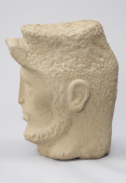 rzeźba; głowa; Komunard - ujęcie z prawej; Głowa męska w czapce od uniformu Gwardii Narodowej z uniesionym daszkiem. Pociągła twarz o delikatnie zaznaczonych rysach uformowana przez minimalistyczne opracowanie jasnego, kamiennego surowca. Nos długi, szyja szeroka. Pozostawione na powierzchni czapki i brody ślady pracy grubym dłutem dają wrażenie miękkości. Zmarszczki ogorzałej skóry mężczyzny uzyskano dzięki pracy cieńszymi dłutami.