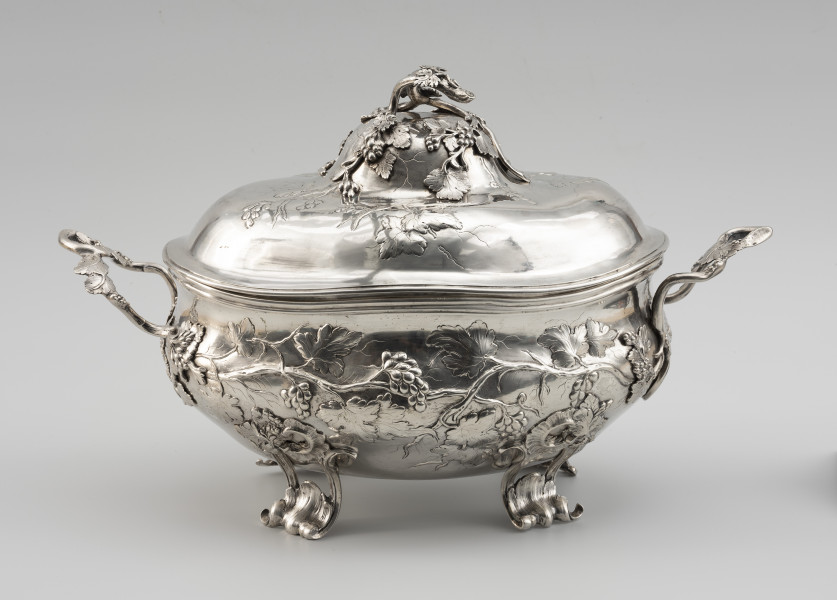 waza - Ujęcie z tyłu; Owalna teryna, wsparta na czterech nóżkach. Korpus o płynnym, organicznym kształcie i falistej linii brzegu, w dolnej części wybrzuszony. Pokrywa wysoka, dwustopniowo wybrzuszona. Zarówno korpus, jak i pokrywa zdobione repusowanymi w płytkim reliefie naturalistycznymi gałązkami winorośli z gronami owoców. Reliefowy ornament miękko przechodzi w wykonane techniką odlewu, plastyczne gałązki winnej latorośli, tworzące dwa uchwyty przy korpusie oraz ozdobne zwieńczenie pokrywy. Nóżki wolutowo wygięte, ażurowe, zdobione rocaillami i gałązkami winorośli.