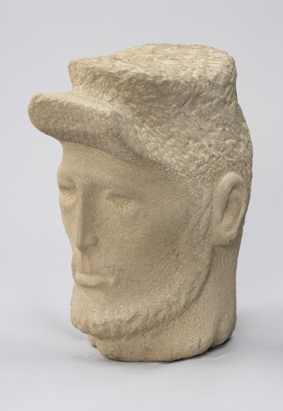 rzeźba; głowa; Komunard - ujęcie ze skosu z prawej; Głowa męska w czapce od uniformu Gwardii Narodowej z uniesionym daszkiem. Pociągła twarz o delikatnie zaznaczonych rysach uformowana przez minimalistyczne opracowanie jasnego, kamiennego surowca. Jedno oko przymrużone, nos długi i szczupły, wokół ust o grubej dolnej wardze delikatny wąsik, zarost zaznaczony też wzdłuż żuchwy, szyja szeroka. Pozostawione na powierzchni czapki i brody ślady pracy grubym dłutem dają wrażenie miękkości. Zmarszczki ogorzałej skóry mężczyzny uzyskano dzięki pracy cieńszymi dłutami.