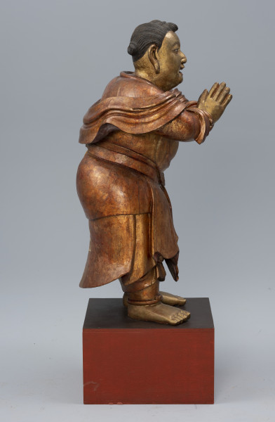 pielgrzym Shancai Tongzi (?) wraz z wypełnieniem - ujęcie z boku; Monumentalna, drewniana, złocona rzeźba postaci męskiej w pozie adoracji przedstawia prawdopodobnie młodego pielgrzyma. Postać stoi na postumencie z rękami wyciągniętymi przed siebie i dłońmi złożonymi w pełnym szacunku geście. Gładko zaczesane czarne włosy zwijają się w niewielkie koczki: z przodu jeden i z tyłu dwa. Pielgrzym ubrany jest w długie, kilkowarstwowe, sięgające za kolana szaty, natomiast z jego ramion i rąk opada tkanina w formie szala. Na jego obu nadgarstkach i kostkach widoczne są bransolety. Stopy ma nagie.