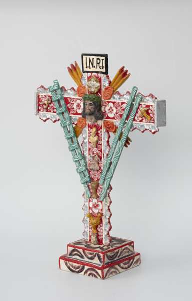 krzyż pasyjny - Ujęcie ze skosu z prawej; Drewniany, czerwony krzyż pasyjny, usytuowany na białej, dwustopniowej, kwadratowej podstawie ozdobionej ciemnym malowanym wzorem półkolistych form i liści, której krawędzie pokryto czerwoną farbą. Bogato dekorowany białymi i fioletowymi wzorami kwiatowymi i obłymi liniami, dodatkowo boki ramion krzyża zostały ozdobione plastycznym falistym motywem w kolorze srebrnym, którego krawędzie podkreślono czerwoną farbą. W jego centralnej części umieszczono wykonaną z formy głowę cierpiącego Chrystusa w koronie cierniowej, nad nią zaś postać gołębia i napisane na prostokątnej plakietce czarnym tuszem litery I.N.R.I. Całość wypełniono rozlicznymi atrybutami i symbolami męki pańskiej, takimi jak: serce, tunika, kielich, kogut, zielona drabina, zielone włócznie (?), szczypce i młotek. Narożniki między skrzyżowanymi ramionami krucyfiksu udekorowano czterema koralowymi różami oraz czterema potrójnymi promieniami w kolorze pomarańczowym.