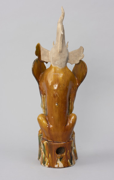 Zhenmushou (strażnik grobowca) - ujęcie z tyłu; Duża, wykonana z porcelany postać. Ciało postaci i podstawa pokryte szkliwem w odcieniach brązu, zieleni, żółci, beżu. Nie zachowały się szkliwienia na głowie, jedynie żółto-zielony fragment na lewym uchu. Z czubka głowy wyrasta stożkowaty róg. Za dużym rogiem znajduje się uszkodzona, nieszkliwiona część o kielichowatym kształcie. Uszy postaci są duże i odstające. Głowę osadzono na torsie uskrzydlonego lwa z kopytami byka. Bestia wydaje się wpół siedzieć i wpół kucać. W podstawie znajduje się okrągły otwór. Pierwotnie brakowało fragmentu prawego skrzydła, małego, środkowego różka na czole oraz zakończenia dużego rogu. Brakujące części uzupełniono podczas prac konserwatorskich.