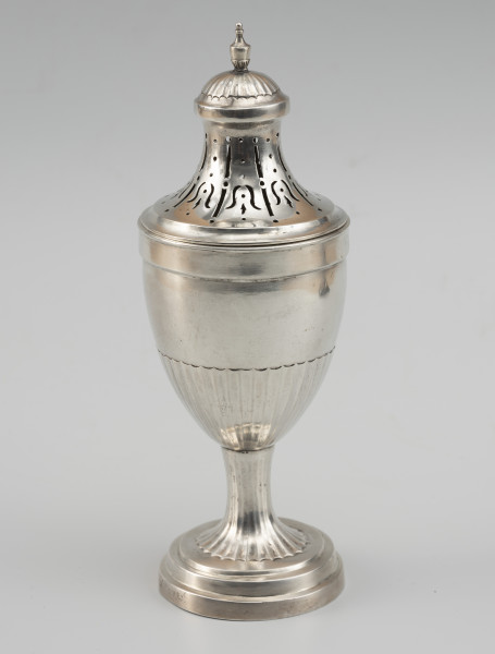 cukiernica do cukru pudru - Ujęcie z przodu; Puszka z ażurową pokrywą, kształtem nawiązująca do antycznych urn. Wsparta na okrągłej stopie z trzema uskokami, przechodzącej w niewysoki, drobno kanelowany trzon. Korpus smukły, o kielichowej formie, w dolnej części również kanelowany. Pokrywa wysoka, o wklęsłych ściankach zakończona półkolistą kopułką, ze zwieńczeniem w formie maleńkiej wazy. Wycięcia na pokrywie układają się we wzór stylizowanych kampanul.