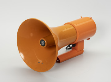 megafon strajkowy - Ujęcie ze skosu z prawej; Metalowy megafon o stożkowatym kształcie fabrycznie pokryty pomarańczową emalią z plastikową obudową i rączką w tym samym kolorze. Rączka w formie otwartej rurki, która mogła służyć do osadzenia głośnika na maszcie lub trzonku odpowiedniej średnicy. Na wewnętrznym stożku tuby głośnika oznakowanie producenta. Z tyłu czarny kabel z poszarpanym końcem, prawdopodobnie pierwotnie zakończony wtyczką, którą można było podłączyć do mikrofonu.