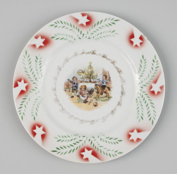 talerz bożonarodzeniowy - Ujęcie z góry; Okrągły płaski talerz z fajansu zdobiony naszkliwnie motywem bożonarodzeniowym. Lustro talerza zdobi kolorowa scena z trójką uśmiechniętych dzieci, bawiących się przy świątecznie przystrojonej, ustawionej na stole choince: chłopiec z lewej ma w ręku pajacyka-marionetkę, dziewczynka siedzi na stołku w centrum sceny z lalka na kolanach, a za nią stoi wózek, na podłodze przed nią leży trąbka i piłka, w prawej części przedstawienia chłopiec siedzi na koniu na biegunach. Przedstawienie okala ażurowy motyw geometryczny, pierwotnie złocony. Kołnierz talerza zdobi kompozycja z malowanych gałązek choinkowych oraz czerwonych gwiazd betlejemskich.