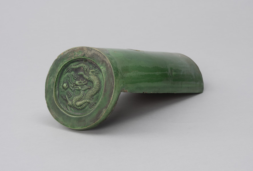 gąsior początkowy ozdobny - Ujęcie ze skosu; Półcylindryczna dachówka typu gąsior z zewnątrz szkliwiona zieloną glazurą. Zasklepiona okrągłą, ozdobą plakietką z odciśniętym, reliefowym wizerunkiem smoka. Smok zieje ogniem, ma rogi, długie wąsy, grzywę, cztery łapy, wijące się, długie, łuskowate ciało. Obok jego paszczy widać płonącą perłę. W plakietce, nad smokiem znajduje się mały, okrągły otwór. Na dachówce widoczne przetarcia, rysy, ubytki, spękania oraz odbarwienia - największe w górnej części plakietki i na grzbiecie – u dołu, przy krawędziach.