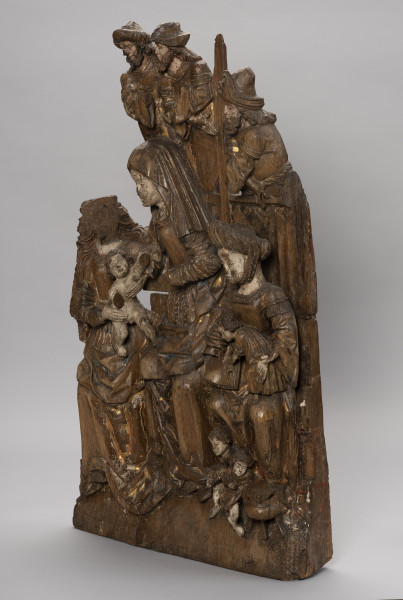 płaskorzeźba ołtarzowa; Rodzina Marii - Ujęcie ze skosu z prawej; Wysoka płaskorzeźbiona płyta z przedstawieniem wielofigurowym, polichromowana. Ukazuje grupę strojnie ubranych kobiet siedzących na ławie i piastujących małe dzieci oraz mężczyzn widocznych powyżej wysokiego zaplecka ławy. W scenie dominuje przedstawienie św. Anny o nieco zwiększonych proporcjach. Święta w chuście na głowie i z różańcem przypiętym do paska wraz z Marią o długich falujących włosach podtrzymują nagiego Jezusa balansującego pomiędzy ich kolanami. Po prawej siedzi Maria Kleofasowa w wysokim marszczonym czepcu z czwórką synów ubranych w tuniki, z których jeden, stojący na kolanach, bawi się otwartą księgą, pozostali biegają wokół kociołka u stóp matki. Brodaci mężczyźni usytuowani za ławą, powyżej kobiet, widoczni w półpostaci, noszą tuniki na koszulach o szerokich rękawach i kunsztownie kształtowane kapelusze. Marszczone suknie z wąskim gorsetem i dekoltami w karo czy obszycia rękawów dekoracyjnymi taśmami, szerokie rękawy koszul to elementy strojów zgodne z niemiecką modą mieszczańską z początku XVI wieku. Modne stroje, łagodne wizerunki okrągłych twarzy a także poruszone pozy i gesty postaci nadają przedstawieniu charakter rodzajowy. Płaskorzeźba wybrana z łączonych bloków drewna, w dolnej części drążona. Płyta częściowo uszkodzona i popękana; brakuje postaci Marii Salome z dziećmi i mężem, męskich wizerunków powyżej Marii a także jej twarzy oraz czwartego dzieciątka u stóp Marii Salome. Polichromia zachowana szczątkowo.