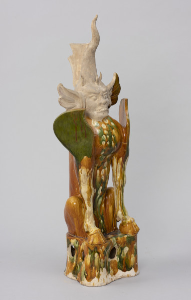 Zhenmushou (strażnik grobowca) - ujęcie ze skosu z lewej; Duża, wykonana z porcelany postać z ludzką twarzą. Ciało postaci i podstawa pokryte szkliwem w odcieniach brązu, zieleni, żółci, beżu. Nie zachowały się szkliwienia na głowie, jedynie żółto-zielony fragment na lewym uchu. Z czubka głowy wyrasta stożkowaty róg, natomiast nad czołem zaznaczono trzy malutkie różki. Za dużym rogiem znajduje się uszkodzona, nieszkliwiona część o kielichowatym kształcie. Uszy postaci są duże i odstające. Głowę osadzono na torsie uskrzydlonego lwa z kopytami byka. Bestia wydaje się wpół siedzieć i wpół kucać. W podstawie znajdują się okrągłe otwory: widoczny jeden od frontu i z boku. Pierwotnie brakowało fragmentu prawego skrzydła, małego, środkowego różka na czole oraz zakończenia dużego rogu. Brakujące części uzupełniono podczas prac konserwatorskich.