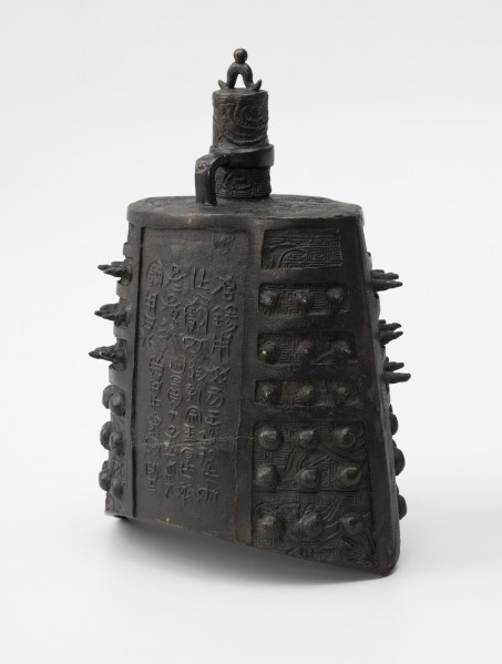 zhong (dzwon chiński) - ujęcie tyłu ze skosu z prawej; Wykonany z brązu dzwon o soczewkowatym przekroju poprzecznym. Na płaskim hełmie znajduje się korona w formie trzpienia, zdobionego rysunkami ptaków, z małym pałąkowatym uszkiem na szczycie zwieńczonym kuleczką i drugim, wygiętym pod kątem prostym, wychodzącym z hełmu i połączonym z trzpieniem za pomocą okrągłej obejmy. Na bogaty ornament reliefowy w postaci siatki wzoru geometrycznego (meander) i zoomorficznego (przedstawienia wężowatych stworzeń, być może smoków) po bokach nałożono kolce mei (w górnej części) oraz guzki (w dolnej części), natomiast w centrum  płaszcza wykonano inskrypcję chińskim pismem małopieczęciowym. Dzwon tego typu nie posiada serca, jego środek pozostaje pusty.