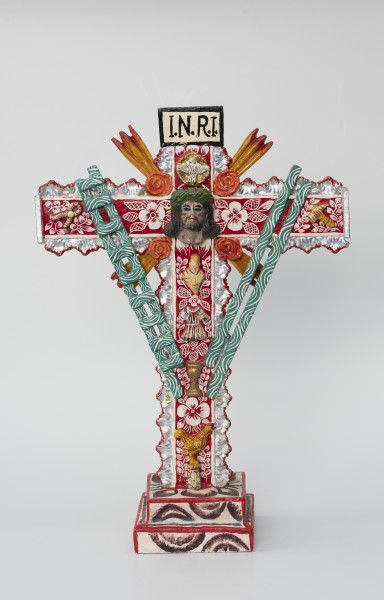 krzyż pasyjny - Ujęcie z przodu; Drewniany, czerwony krzyż pasyjny, usytuowany na białej, dwustopniowej, kwadratowej podstawie ozdobionej ciemnym malowanym wzorem półkolistych form i liści, której krawędzie pokryto czerwoną farbą. Bogato dekorowany białymi i fioletowymi wzorami kwiatowymi i obłymi liniami, dodatkowo boki ramion krzyża zostały ozdobione plastycznym falistym motywem w kolorze srebrnym, którego krawędzie podkreślono czerwoną farbą. W jego centralnej części umieszczono wykonaną z formy głowę cierpiącego Chrystusa w koronie cierniowej, nad nią zaś postać gołębia i napisane na prostokątnej plakietce czarnym tuszem litery I.N.R.I. Całość wypełniono rozlicznymi atrybutami i symbolami męki pańskiej, takimi jak: serce, tunika, kielich, kogut, zielona drabina, zielone włócznie (?), szczypce i młotek. Narożniki między skrzyżowanymi ramionami krucyfiksu udekorowano czterema koralowymi różami oraz czterema potrójnymi promieniami w kolorze pomarańczowym.