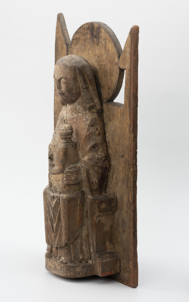 Tronująca Madonna z Dzieciątkiem - Ujęcie ze skosu z prawej; Niewielka figura tronującej Marii z dzieciątkiem o zwartej bryle, przytwierdzona do wysokiego zaplecka o geometrycznym zwieńczeniu, polichromowana. Rzeźba w typie Sedes Sapientiae ukazuje frontalnie siedzącą na tronie Marię w sztywnej, wyprostowanej pozie z dzieckiem w wieku chłopięcym usytuowanym bokiem, w pozycji siedzącej, ponad jej lewym kolanem. Długa suknia i płaszcz Marii oraz tunika Jezusa gładko przylegają do ciał postaci tworząc nieznaczne, schematyczne draperie w partii nóg. Między goleniami Marii tkanina układa się w charakterystyczne trójkątne fałdy o ostrych krawędziach. Hieratyczne pozy postaci przełamuje gest dłoni matki przytrzymującej syna. Głowa Marii osadzona na sztywnej, szerokiej szyi okryta chustą ułożoną w tylnej partii na kształt welonu sięgającego do ramion. Twarz pociągła, owalna, zastygła w pozbawionym emocji wyrazie. Symetryczna fryzura chłopca z falujących włosów sięga poniżej uszu. Zwieńczenie gładko opracowanego zaplecka tworzy kolisty nimb za głową Marii flankowany trójkątnymi sterczynami oparcia tronu. Rzeźba opracowana jako przyścienna. Brak prawej dłoni Marii. Powierzchniowe ubytki drewna i otwory po drewnojadach. Polichromia zachowana szczątkowo.