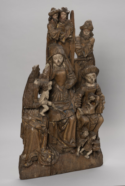 płaskorzeźba ołtarzowa; Rodzina Marii - Ujęcie ze skosu z lewej; Wysoka płaskorzeźbiona płyta z przedstawieniem wielofigurowym, polichromowana. Ukazuje grupę strojnie ubranych kobiet siedzących na ławie i piastujących małe dzieci oraz mężczyzn widocznych powyżej wysokiego zaplecka ławy. W scenie dominuje przedstawienie św. Anny o nieco zwiększonych proporcjach. Święta w chuście na głowie i z różańcem przypiętym do paska wraz z Marią o długich falujących włosach podtrzymują nagiego Jezusa balansującego pomiędzy ich kolanami. Po prawej siedzi Maria Kleofasowa w wysokim marszczonym czepcu z czwórką synów ubranych w tuniki, z których jeden, stojący na kolanach, bawi się otwartą księgą, pozostali biegają wokół kociołka u stóp matki. Brodaci mężczyźni usytuowani za ławą, powyżej kobiet, widoczni w półpostaci, noszą tuniki na koszulach o szerokich rękawach i kunsztownie kształtowane kapelusze. Marszczone suknie z wąskim gorsetem i dekoltami w karo czy obszycia rękawów dekoracyjnymi taśmami, szerokie rękawy koszul to elementy strojów zgodne z niemiecką modą mieszczańską z początku XVI wieku. Modne stroje, łagodne wizerunki okrągłych twarzy a także poruszone pozy i gesty postaci nadają przedstawieniu charakter rodzajowy. Płaskorzeźba wybrana z łączonych bloków drewna, w dolnej części drążona. Płyta częściowo uszkodzona i popękana; brakuje postaci Marii Salome z dziećmi i mężem, męskich wizerunków powyżej Marii a także jej twarzy oraz czwartego dzieciątka u stóp Marii Salome. Polichromia zachowana szczątkowo.